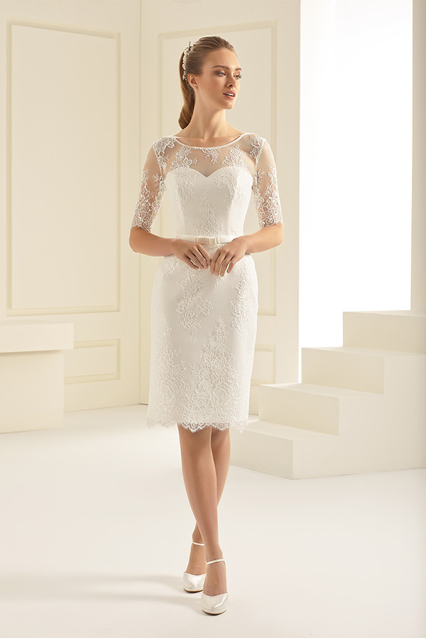 Junge Frau mit blondem Haar trägt ein kurzes weißes Spitzenkleid und blickt ruhig