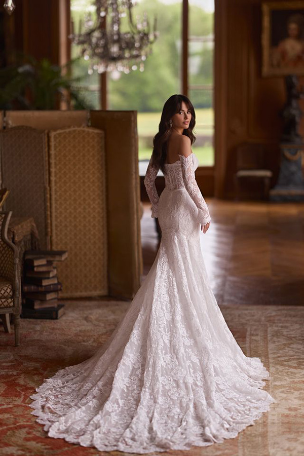 Eine Frau mit langen dunklen Haaren trägt ein edles Spitzen-Brautkleid und blickt elegant zurück