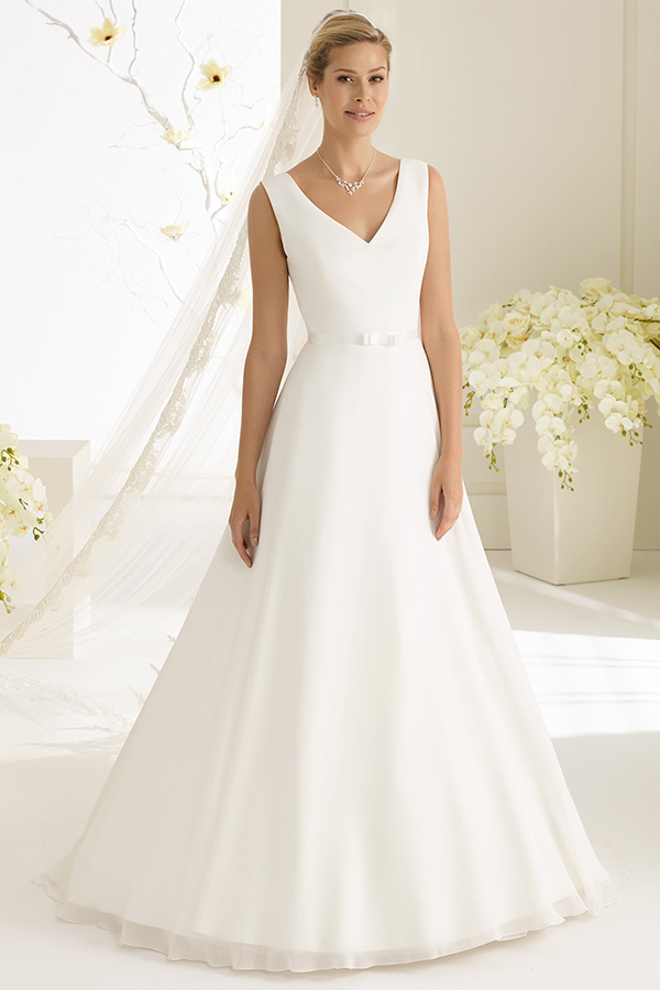 Eine blonde Frau in weißem Brautkleid lächelt dezent vor hellen Blumenarrangements