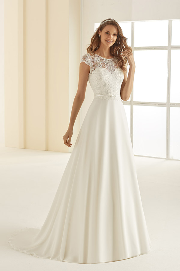 Eine lächelnde Frau in weißem Brautkleid mit Spitzenoberteil steht vor Fenstern