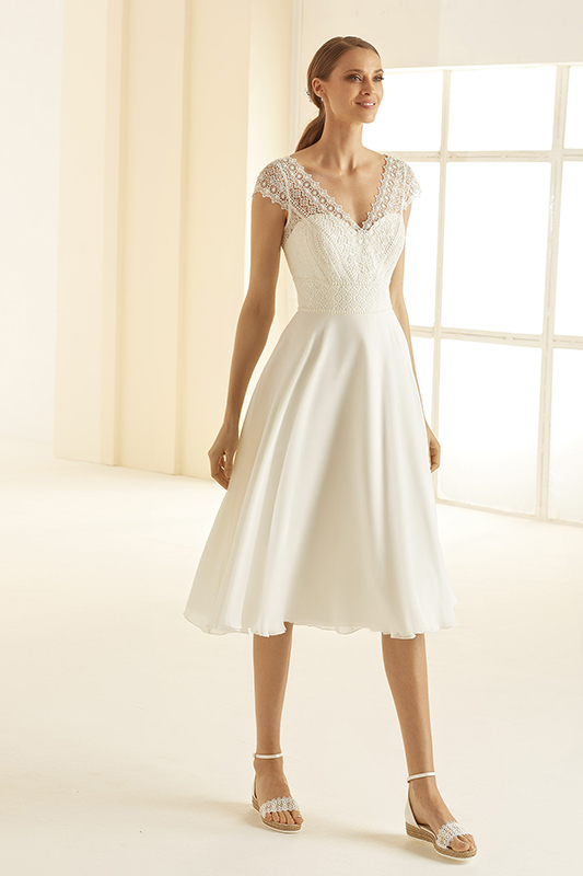 Eine lächelnde Frau im weißen Spitzenkleid mit Perlendetails und schimmernden Sandalen