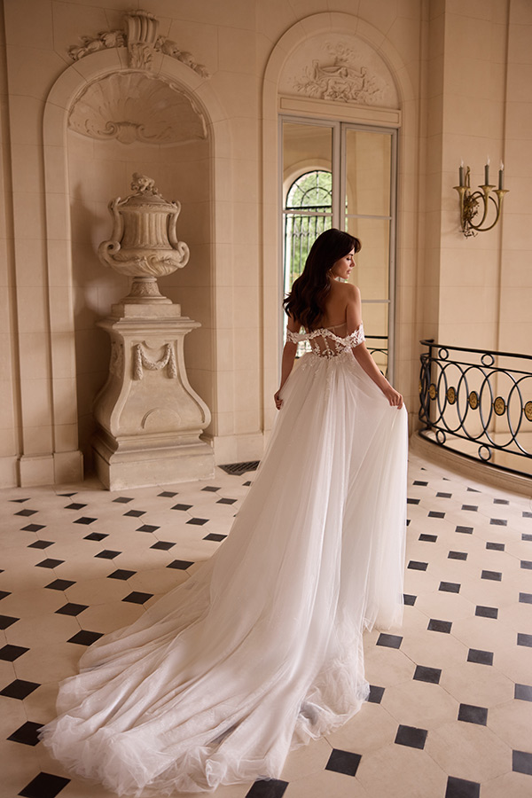 Eine brünette Frau im eleganten weißen Brautkleid steht mit sanftem Lächeln in einem klassischen, hellen Saal mit großen Fenstern und schwarz-weiß gefliestem Boden