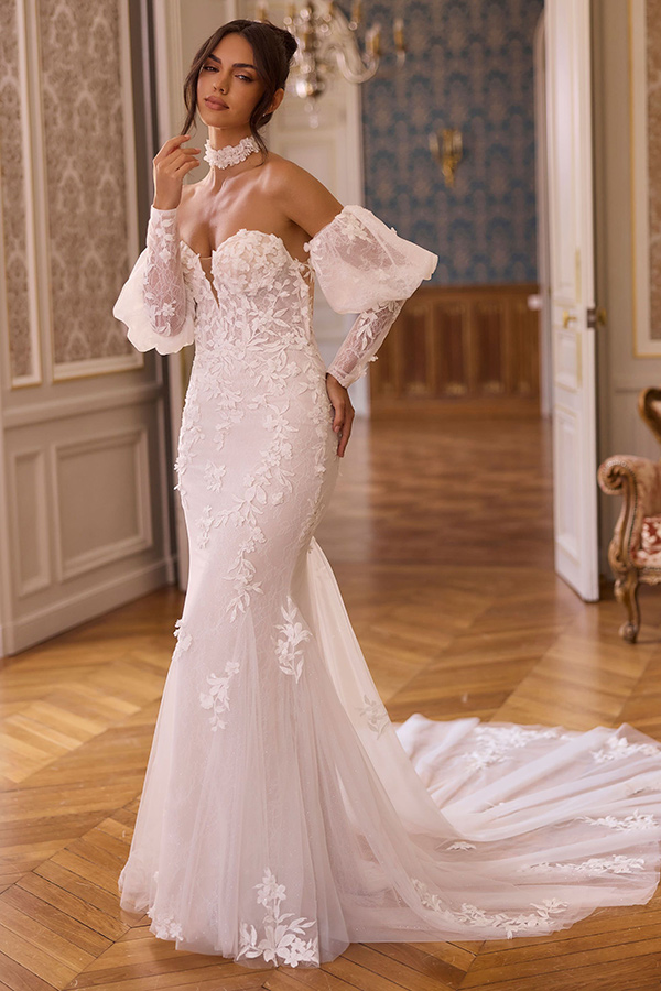 Eine dunkelhaarige Frau im eleganten weißen Spitzenkleid steht ernst im edlen Saal