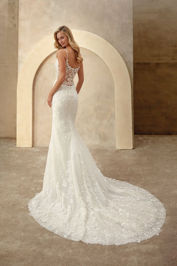 Eine blonde Frau blickt lächelnd über die Schulter und trägt ein elegantes weißes Spitzenkleid mit langem Schleppe vor beigem Hintergrund