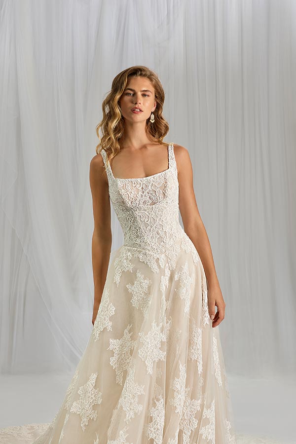 Eine junge Frau mit lockigem Haar lächelt leicht und trägt ein elegantes, cremefarbenes Brautkleid mit Spitzenmuster