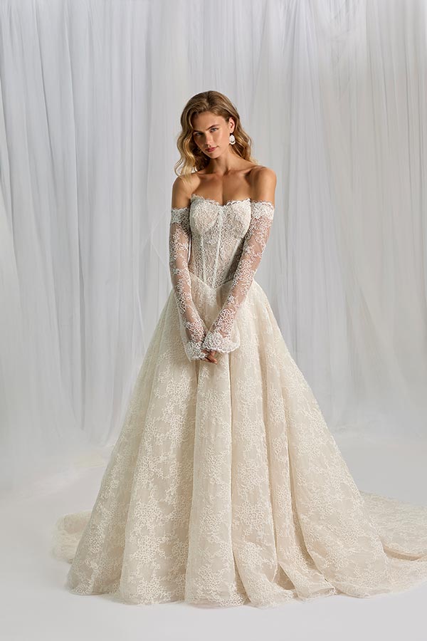 Eine blonde Frau mit sanftem Lächeln trägt ein cremefarbenes Spitzenkleid mit langen Ärmeln