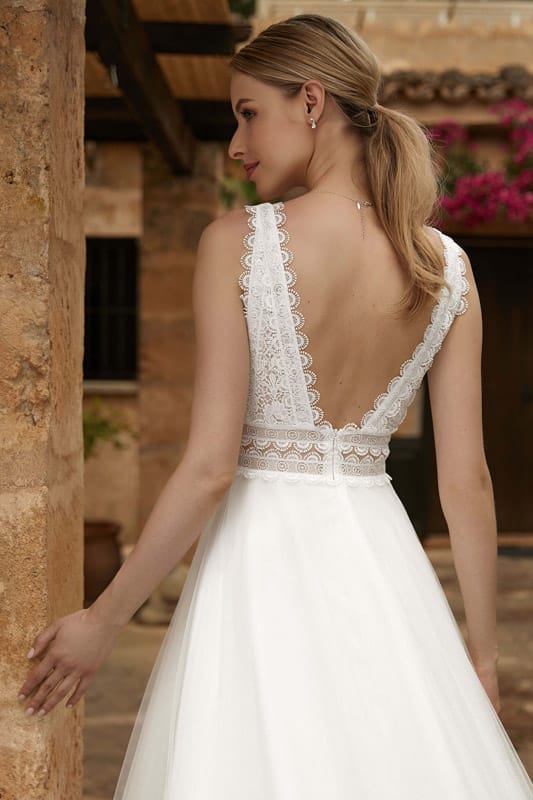 Eine blonde Frau im weißen Brautkleid mit Spitzenrücken blickt lächelnd zur Seite