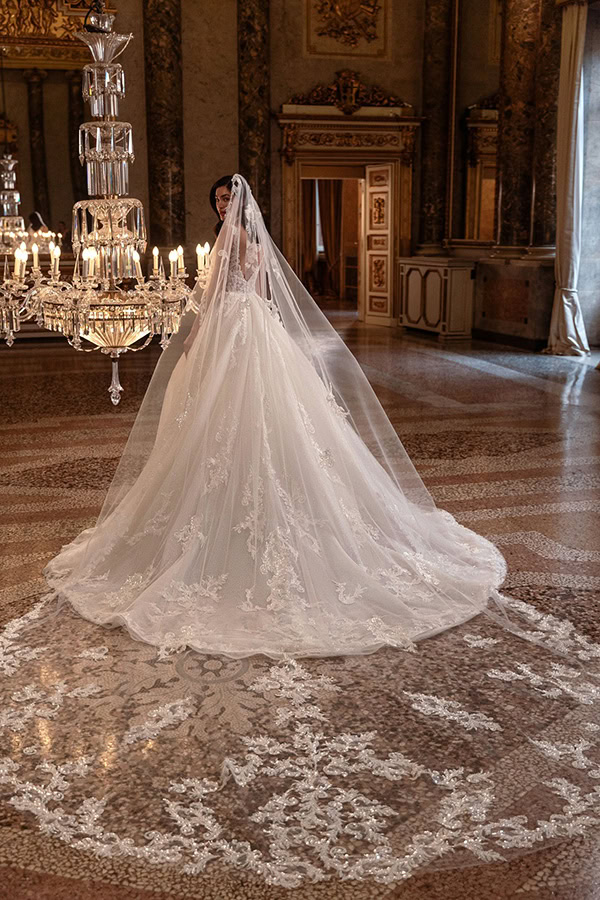 Eine Frau im weißen Brautkleid mit langem Schleier steht in elegantem Saal unter Kronleuchter