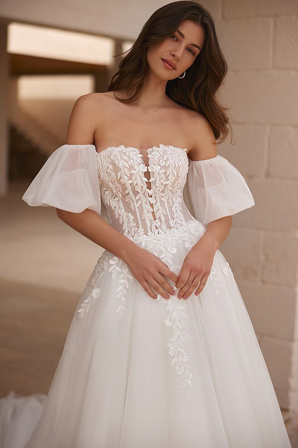 Junge Frau mit langen braunen Haaren trägt ein weißes Spitzenbrautkleid und schaut ruhig