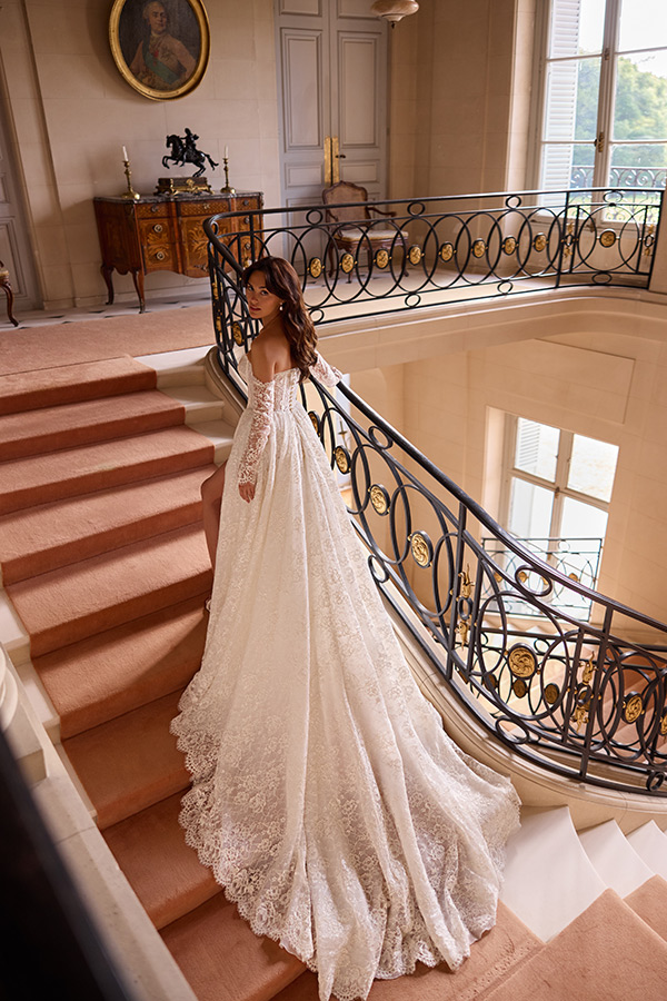 Eine braunhaarige Frau im eleganten weißen Spitzenkleid steht lächelnd auf einer prunkvollen Treppe