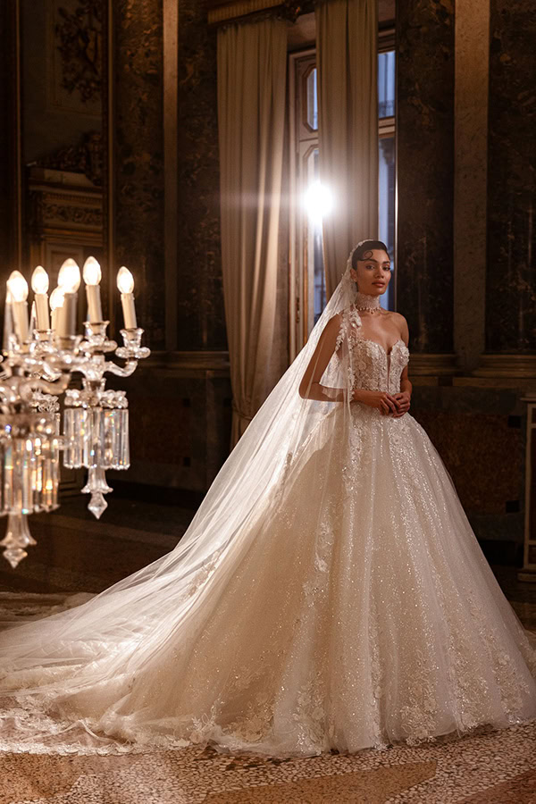 Eine braunhaarige Frau im funkelnden weißen Brautkleid steht elegant in prunkvollem Saal