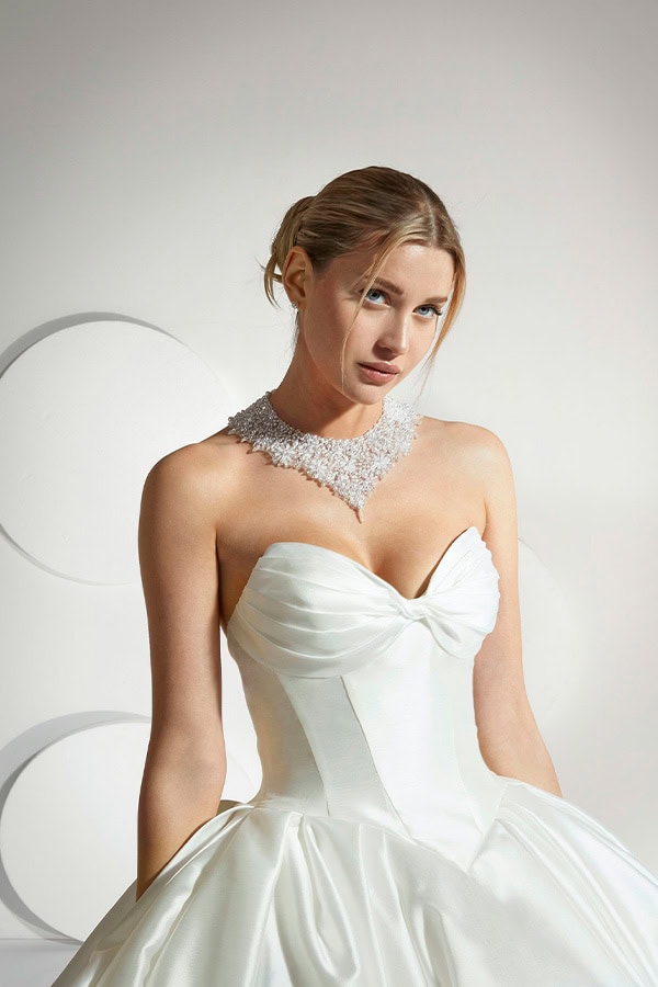 Eine blonde Frau in weißem Brautkleid und glitzernder Halskette blickt ernst