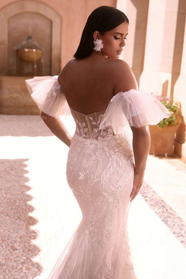 Eine Frau mit langen dunklen Haaren trägt ein elegantes weißes Brautkleid mit floraler Spitze und transparentem Rückenteil, ihr Ausdruck wirkt verträumt