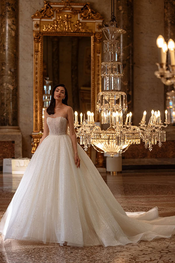 Eine junge Frau im weißen glitzernden Brautkleid steht elegant vor einem großen Spiegel