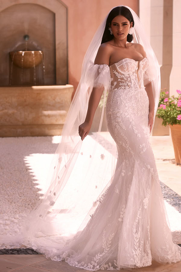 Eine Frau mit ernstem Blick trägt ein schulterfreies Spitzenbrautkleid in Weiß