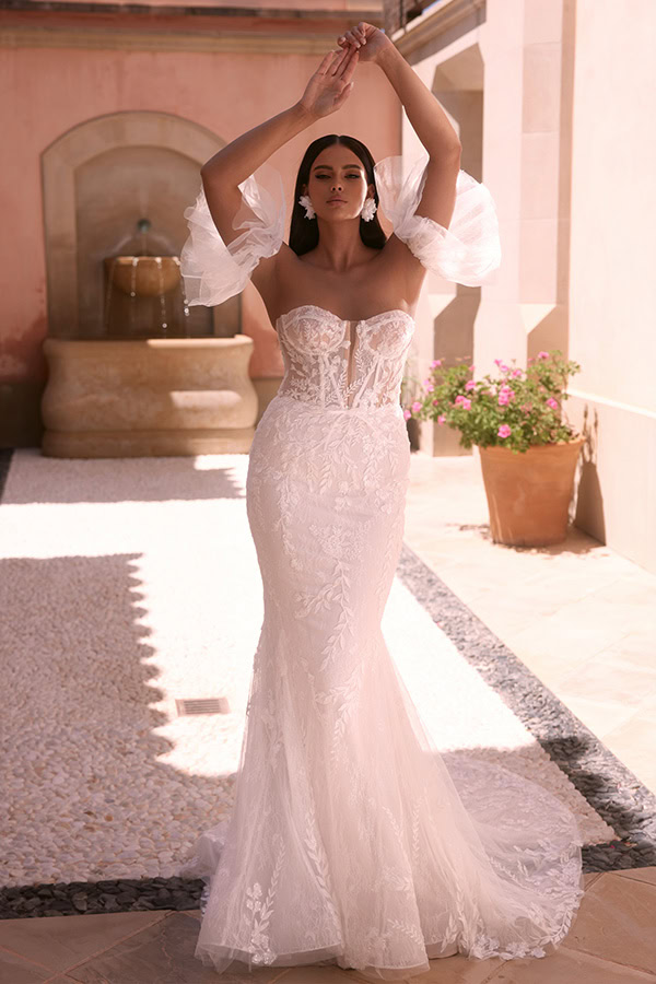 Frau mit neutralem Gesichtsausdruck trägt ein elegantes weißes Spitzenkleid und hebt die Arme