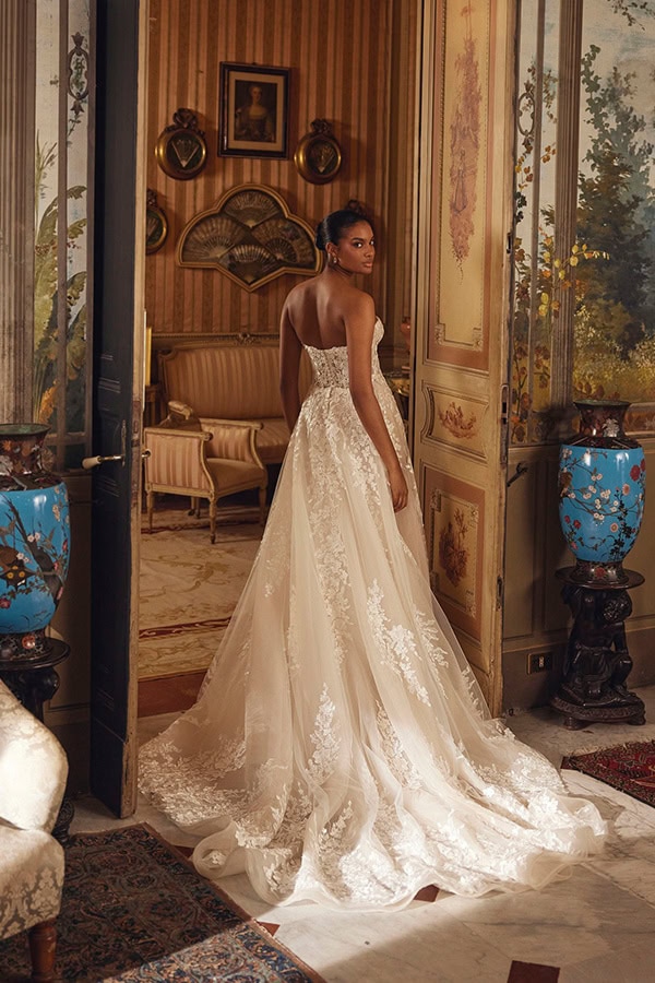 Eine lächelnde Frau im cremefarbenen Spitzenkleid steht elegant in einem prunkvollen Raum