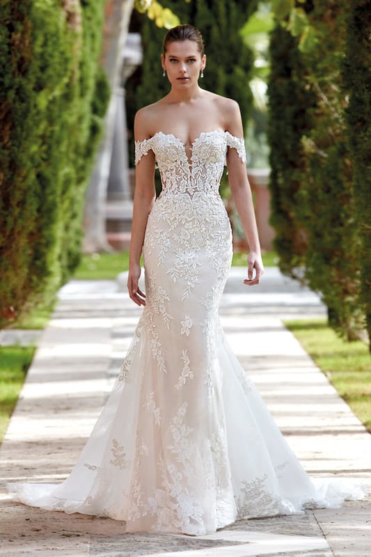 Eine junge Frau mit ernstem Blick trägt ein elegantes weißes Spitzenkleid mit Carmen-Ausschnitt