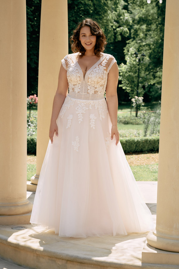 Eine Frau mit lockigem braunen Haar lächelt und trägt ein cremefarbenes Hochzeitskleid
