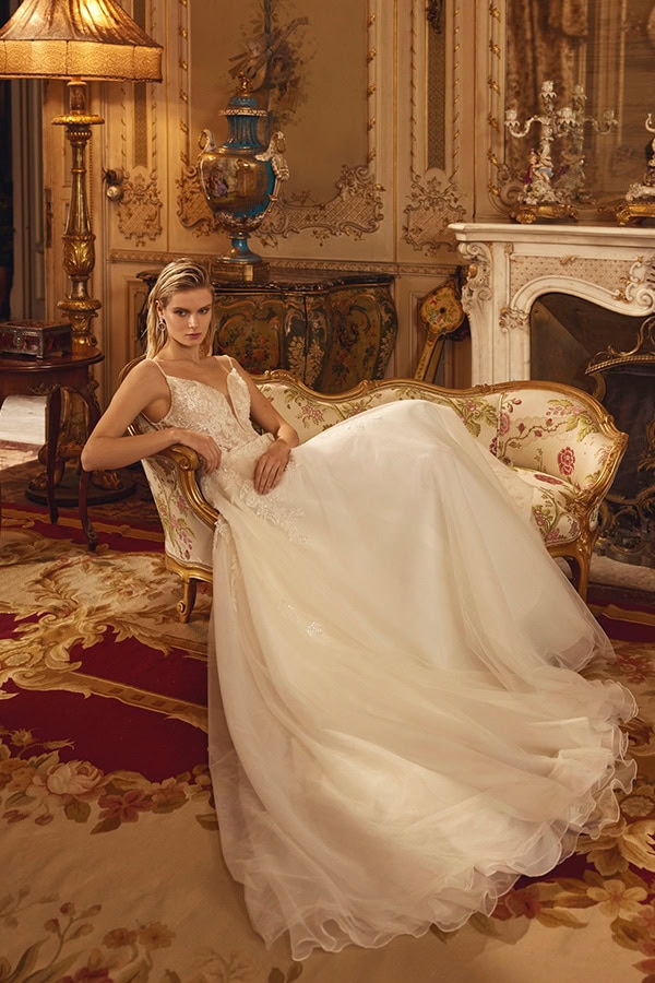Eine junge Frau mit ernster Miene trägt ein weißes Brautkleid auf einem goldenen Sofa im prunkvollen Raum