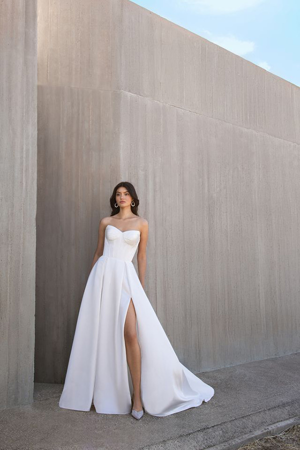 Junge Frau mit ernstem Blick trägt weißes Kleid mit hohem Beinschlitz vor Betonwand