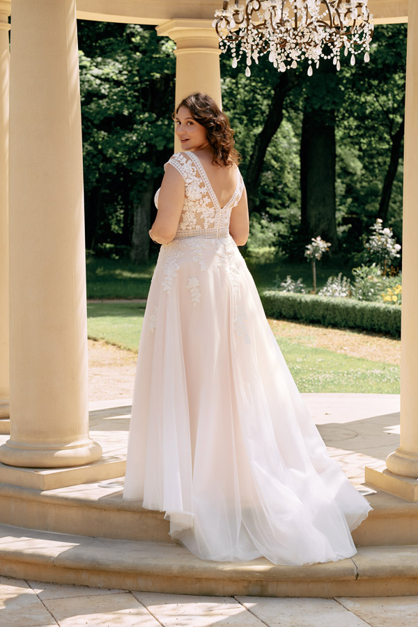 Eine Frau mit lockigem braunem Haar lächelt leicht in einem eleganten weißen Brautkleid unter einem Säulengang