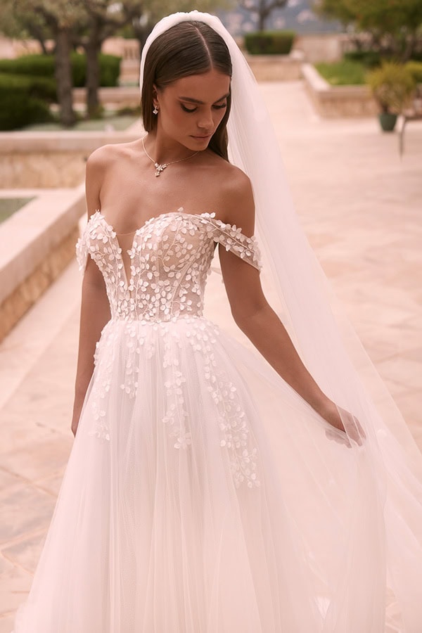 Eine junge Frau mit ernstem Ausdruck trägt ein weißes Brautkleid mit floralen Applikationen und Schleier