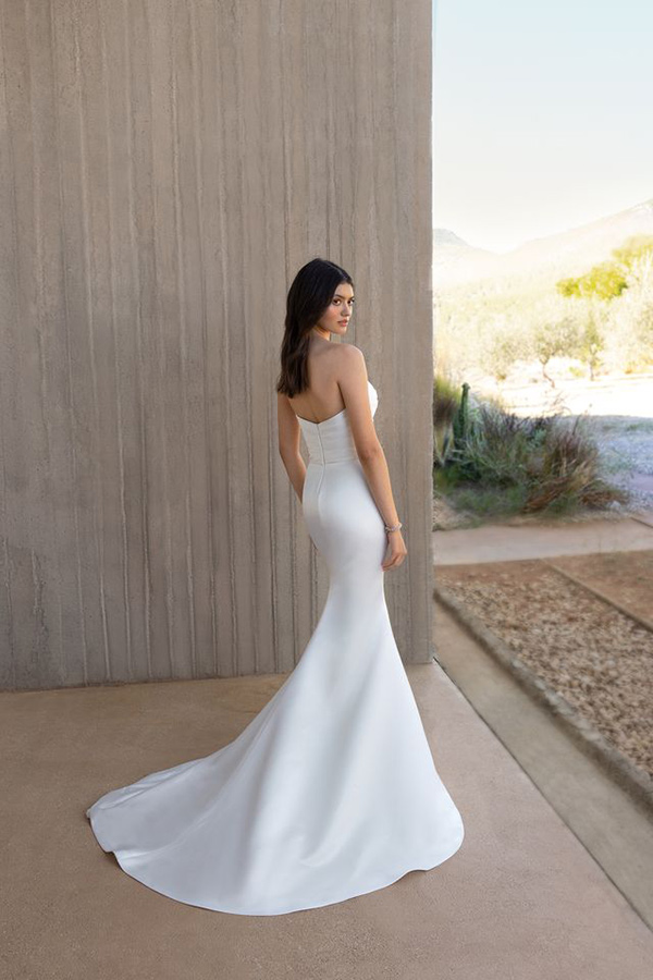 Eine junge Frau mit langen dunklen Haaren trägt ein elegantes weißes Brautkleid und blickt ernst über die Schulter