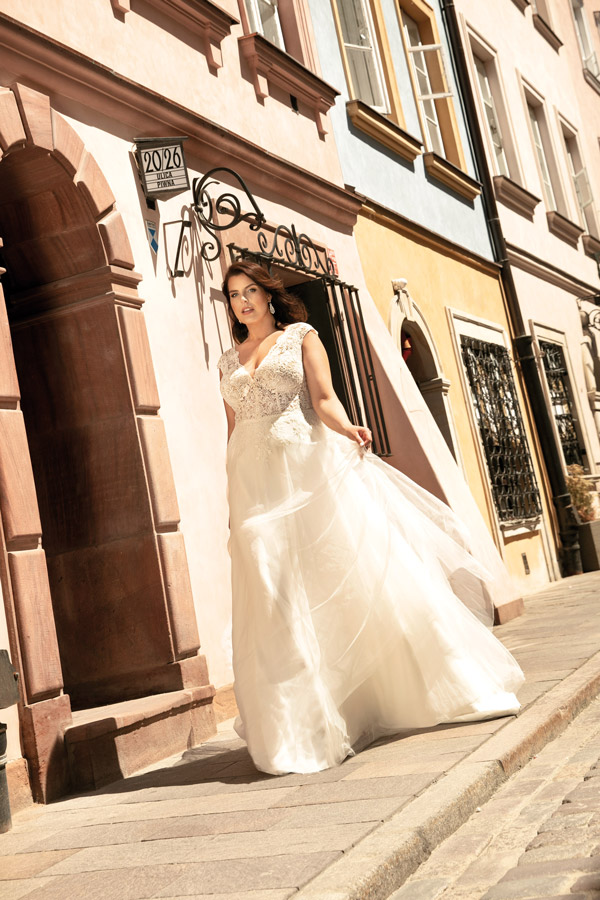 Eine Frau im weißen Brautkleid geht lächelnd durch eine sonnige Altstadtstraße