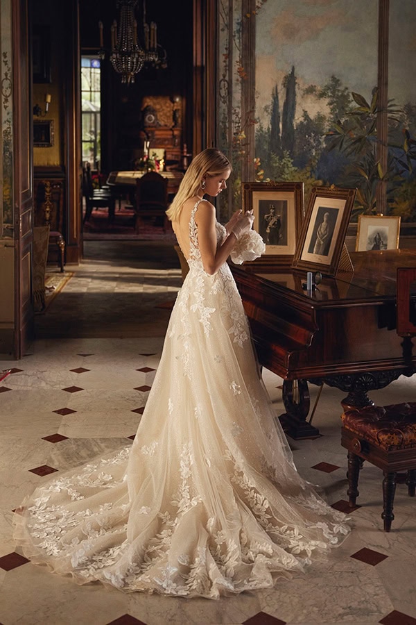 Eine blonde Frau im eleganten Spitzenkleid steht nachdenklich am Flügel im Salon