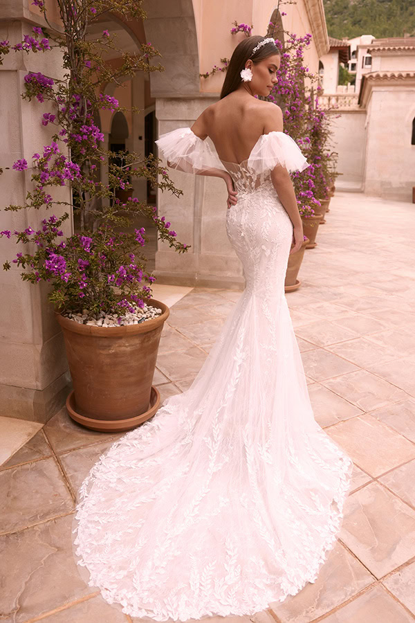 Frau in elegantem weißen Brautkleid mit transparenter Spitze und Blumenmuster steht lächelnd auf einer Terrasse vor violetten Blumentöpfen
