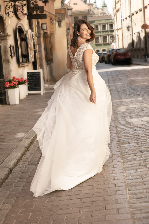 Frau mit lockigem braunen Haar lächelt glücklich im weißen Brautkleid auf Kopfsteinpflasterweg