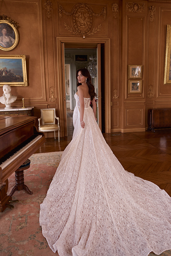 Eine Frau mit langen braunen Haaren steht in einem edlen Raum und trägt ein elegantes cremefarbenes Spitzenkleid mit langer Schleppe