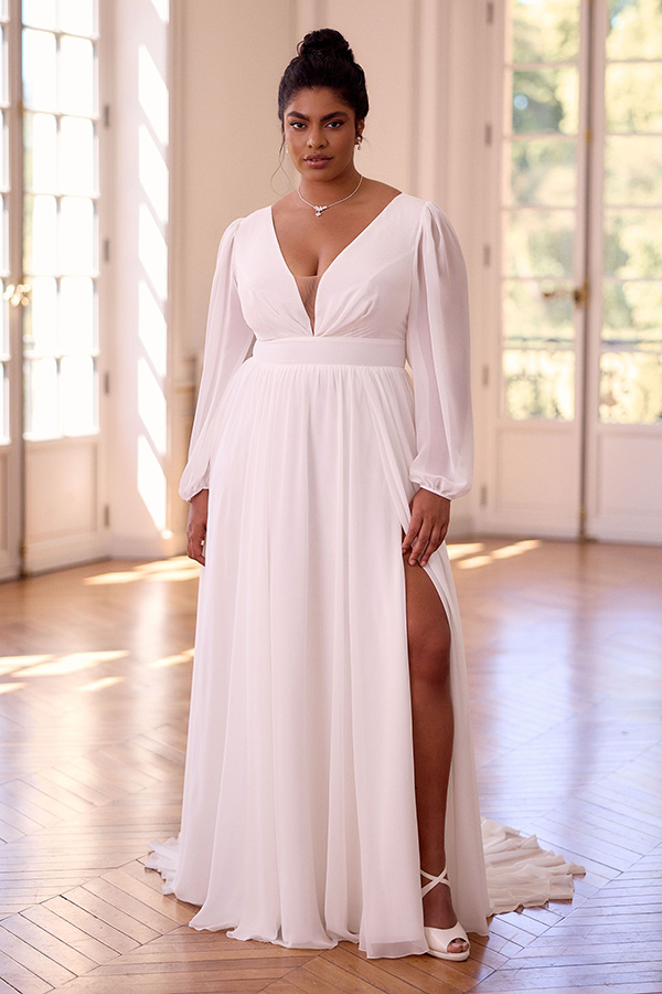 Eine Frau mit ernstem Blick trägt ein weißes Kleid mit tiefem Ausschnitt und Beinschlitz