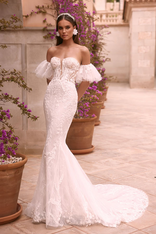 Eine brünette Frau blickt selbstbewusst in einem weißen Spitzenkleid elegant in die Kamera