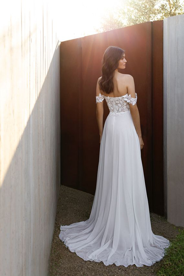 Eine Frau mit dunklem Haar trägt ein weißes, schulterfreies Kleid und steht ruhig draußen