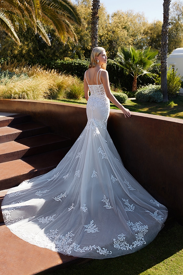 Eine elegante Frau in weißem Brautkleid steht lächelnd im sonnigen Garten