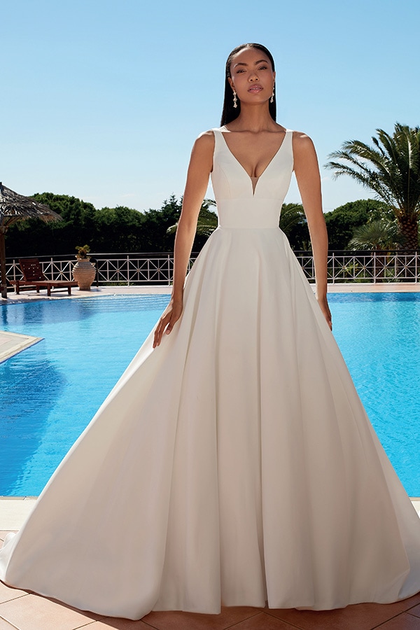 Eine elegant gekleidete Frau mit langem weißen Kleid posiert ernst vor einem Pool