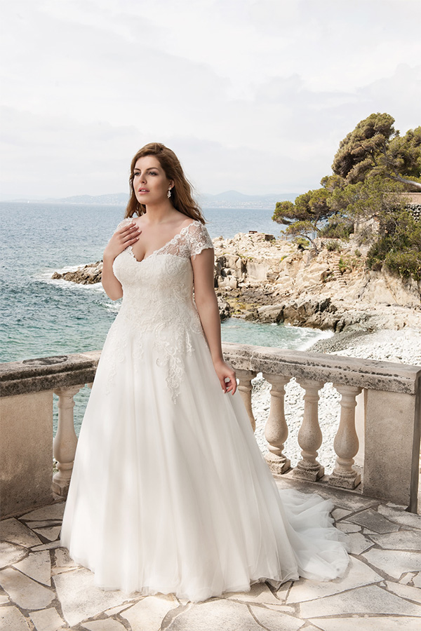 Eine Frau in einem eleganten weißen Brautkleid steht nachdenklich am Meer