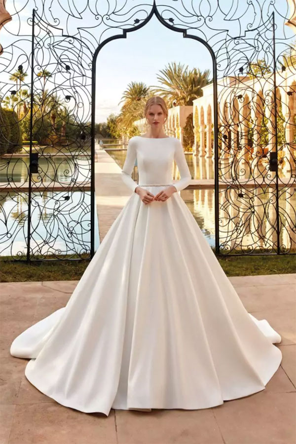 Eine blonde Frau im eleganten weißen Brautkleid steht ernst vor kunstvollem Tor