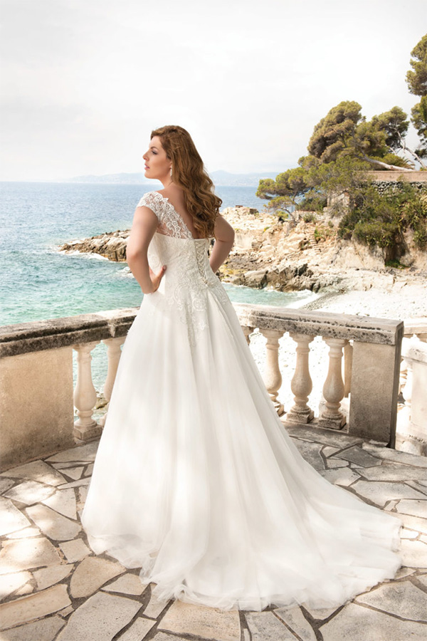 Eine Frau im weißen Brautkleid steht auf einer Terrasse am Meer und blickt verträumt seitlich