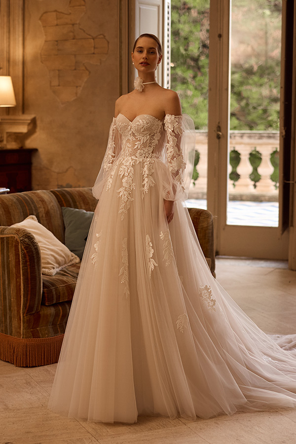 Eine junge Frau in cremefarbenem Brautkleid mit Spitzenärmeln steht ruhig im eleganten Raum