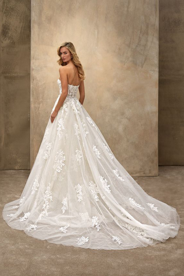 Eine junge Frau mit langen blonden Haaren trägt ein weißes Brautkleid mit Spitzenornamenten und schaut nach hinten