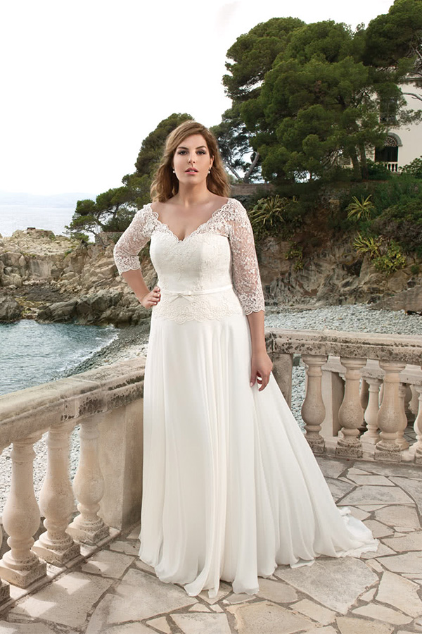 Eine Frau in weißem Hochzeitskleid mit Spitzenärmeln steht ernst am Meer