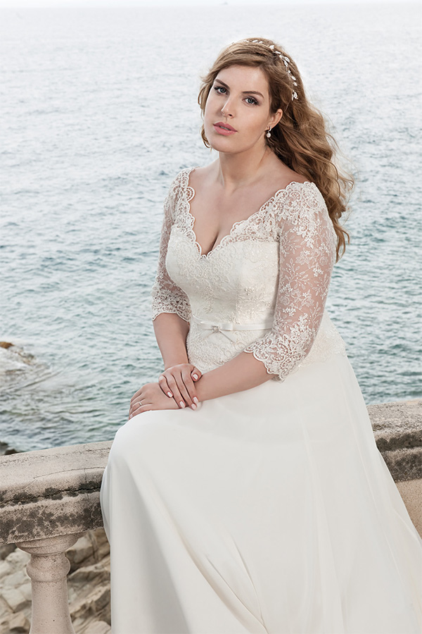 Eine Frau mit leichtem Lächeln trägt ein cremeweißes Spitzenkleid am Wasser