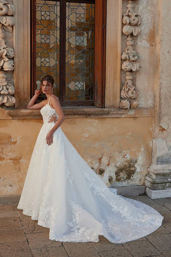 Eine junge Frau im weißen Brautkleid mit Spitzenapplikationen steht vor einem alten Fenster und blickt nachdenklich