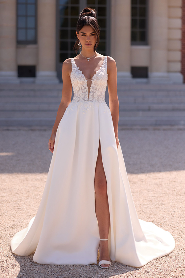 Eine brünette Frau mit ernster Miene trägt ein weißes Brautkleid mit Spitzenbesatz und hohem Beinschlitz vor einem klassischen Gebäude