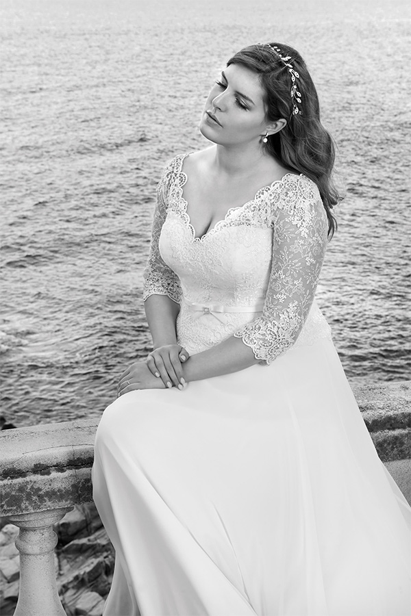 Eine nachdenkliche Frau im eleganten weißen Brautkleid mit Spitzenärmeln sitzt am Meer