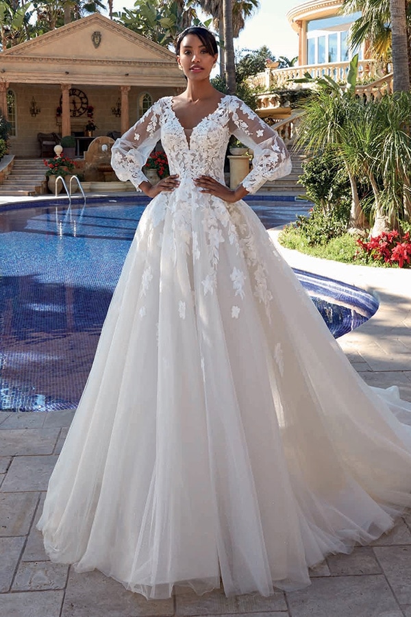 Eine elegante Frau in einem prachtvollen weißen Brautkleid steht vor einem Pool und lächelt leicht