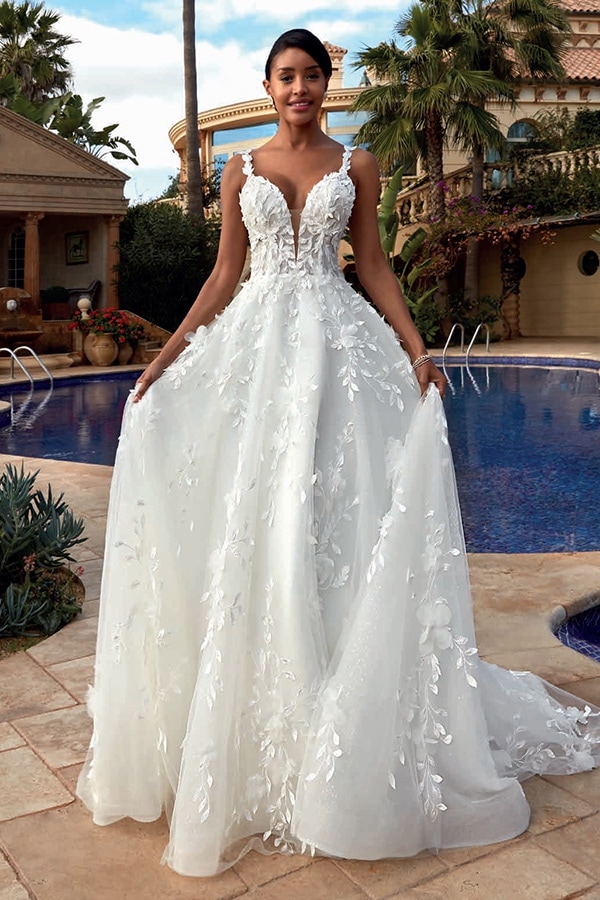 Eine elegant lächelnde Frau im weißen Hochzeitskleid mit floraler Stickerei vor Pool